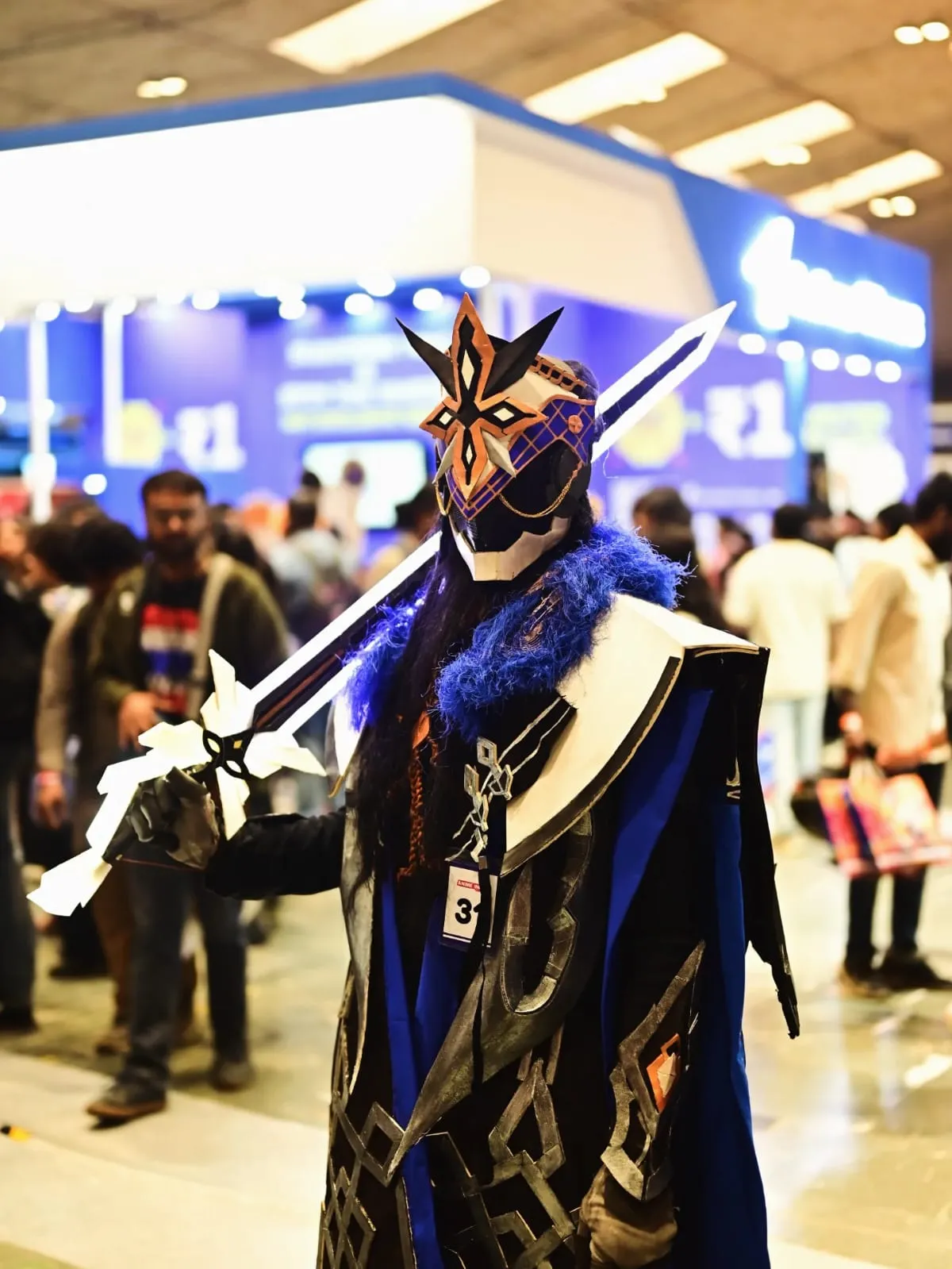 Cosplayer dressed as Il Capitano from Genshin Impact at an anime convention, wielding a greatsword and wearing his signature Fatui Harbinger star mask and blue fur mantle.